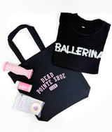 A black 'DEAD POINTE SHOE CLUB' tote bag is laid flat on a white background alongside a folded black T-shirt with 'BALLERINA' in white, a pink Arch Angel, foot massage roller, a Pointe Powder 3oz bottle, and a packet of Toe-Pro Tape.