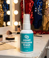 Bottle of 'Covet Costume Fresh SPRITZ' on a dressing room vanity with decorative lights and sequined costumes in the background
