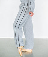 A dancer wearing light heather gray wide-leg sweatpants with black side stripes and the COVET DANCE printed vertically on one leg, paired with ballet pointe shoes against a plain light gray studio background.