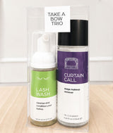 A boxed set of three beauty products labeled 'TAKE A BOW TRIO' on a light wood surface against a white wall. The set includes a green 'LASH WASH' pump bottle, a heart-shaped hair clip, and a purple-and-white 'CURTAIN CALL stage makeup remover' bottle inside a clear plastic package.