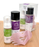 A post-performance set displayed on a light wood surface, including two bottles labeled 'Curtain Call' and 'Lash Wash,' a pink heart-shaped hair clip, and a clear box with a purple label reading 'Take a Bow Trio.'.