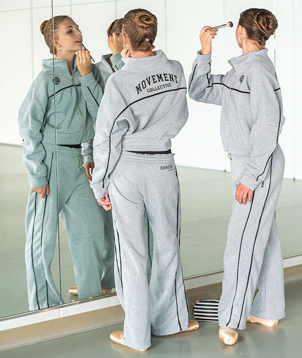 Two dancers in a gray heathered dancewear tracksuit with black piping and 'MOVEMENT COLLECTION' printed on the back, seen from behind while putting on makeup in a mirror.