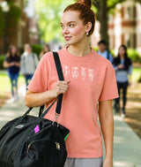 A ballerina walking outdoors on a college campus-style path wearing a coral pink graphic t-shirt with four white pointe shoes graphic, carrying a black dance bag.