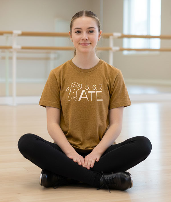 Tap dancer sitting on the floor in a dance studio wearing a gingerbread brown t-shirt with 5, 6, 7, ATE design