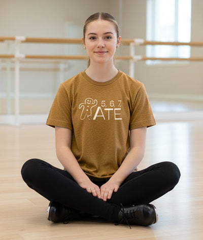 Tap dancer sitting on the floor in a dance studio wearing a gingerbread brown t-shirt with 5, 6, 7, ATE design
