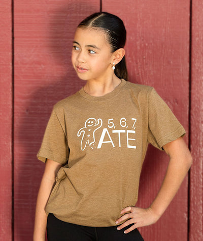 Young dancer wearing a brown t-shirt with a 5, 6, 7, ATE design in front of a red wooden wall.