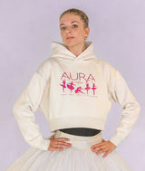 A ballerina modeling a cream-colored cropped hoodie with a pink graphic print that reads 'AURA +1000' and features ballerina silhouettes, paired with a white tutu skirt against a light lavender background.