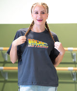 A dance teacher with two braids smiling and wearing a dark gray t-shirt with a colorful 'Back to the Studio' graphic, set against a green and white dance studio background.