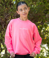A young girl wearing a bright pink crewneck sweatshirt with 'BALLERINA' embroidered in white on the chest, standing outdoors in front of a blurred green foliage background.
