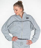A woman wearing a light gray heather zip-up sweatshirt with black piping details and a blackCovet Dance logo on the left chest, paired with matching gray sweatpants against a plain white background.