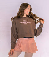 A woman modeling a brown cropped sweatshirt with 'DEAD POINTE SHOE CLUB' printed in pink on the front, paired with a peach ruffled skirt, against a light lavender studio background.