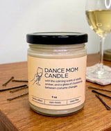 'Dance Mom Candle' jar on a wooden nightstand with bobbypins and a glass of wine in the background.