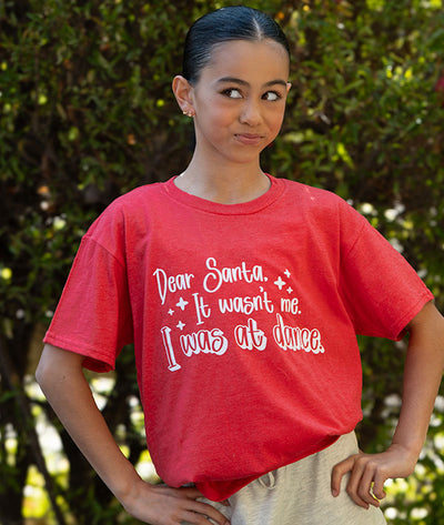 Young dancer wearing a red t-shirt with "Dear Santa, It wasn't me" graphic standing outdoors against a green leafy background