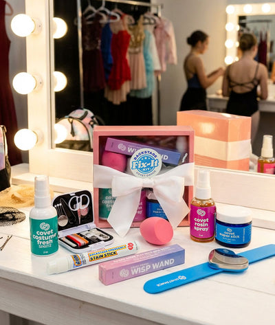 Covet Dance Backstage Fix-It Kit on a dressing room table with dancers getting ready for a performance visible in the mirror and costumes in the background