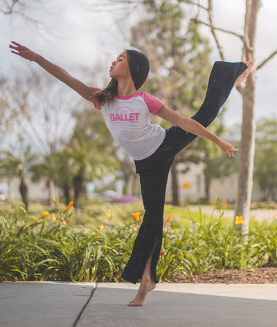 Young ballerina in arabesque wearing Covet Dance raglan tee that says, "Just another day at the barre - BALLET"