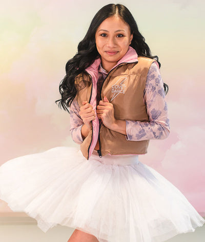 Ballerina wearing a tan puffy vest with 'Covet Dance' logo, pink shirt, and white tutu against a pastel gradient background.