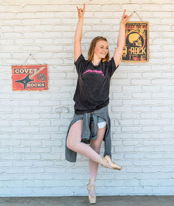 Person wearing a black vintage t-shirt with heavy metal 'Ballerina' design, standing against a white brick wall with vintage rock signs.