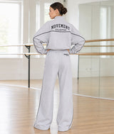A woman stands in a dance studio wearing a light gray two-piece tracksuit from behind, featuring a cropped sweatshirt with 'MOVEMENT COLLECTIVE' printed on the back and matching wide-leg sweatpants.