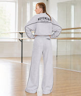 A woman stands with her back to the camera in a dance studio, wearing a light gray sweat jacket and matching sweatpants. The tracksuit features 'MOVEMENT COLLECTIVE' printed in black across the upper back, and the sweatpants have 'DANCER No. 5,678' printed on the back hip.