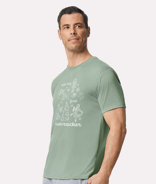 Adult wearing a light green tee shirt that says "Not my first nutcracker" in white print, and depicts the Nutcracker, Mouse King, Clara doing ballet in pointe shoes, and Christmas themed items: snowflakes, candy canes, a drum, a Christmas tree, a wrapped present, and a gingerbread man.