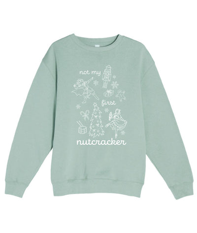 Light green sweatshirt that says "Not my first nutcracker" in white print, and depicts the Nutcracker, Mouse King, Clara doing ballet in pointe shoes, and Christmas themed items: snowflakes, candy canes, a drum, a Christmas tree, a wrapped present, and a gingerbread man.