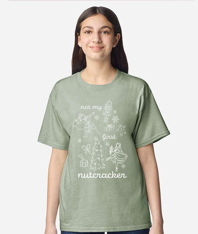 Girl wearing light green tee shirt that says "Not my first nutcracker" in white print, and depicts the Nutcracker, Mouse King, Clara doing ballet in pointe shoes, and Christmas themed items: snowflakes, candy canes, a drum, a Christmas tree, a wrapped present, and a gingerbread man.