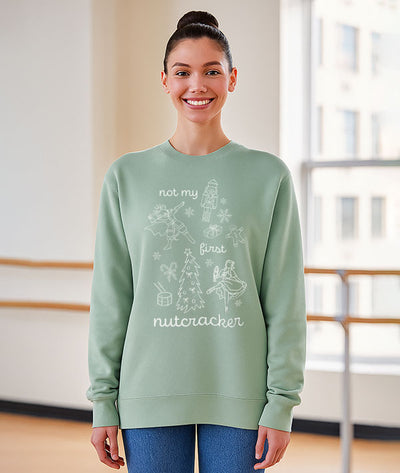 A dancer standing in a dance studio, wearing a light green sweatshirt that says "Not my first nutcracker" in white print, and depicts the Nutcracker, Mouse King, Clara doing ballet in pointe shoes, and Christmas themed items: snowflakes, candy canes, a drum, a Christmas tree, a wrapped present, and a gingerbread man.