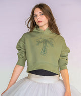 A woman wearing a cropped sage green hoodie with a holly mistletoe garland and ballet pointe shoe graphic and text on the chest, styled with a white tutu skirt, against a light lavender studio background.