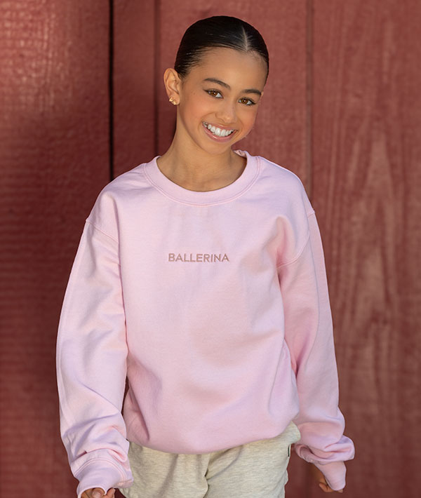 Dancer wearing an embroidered pink 'BALLERINA' sweatshirt against a red-brown background
