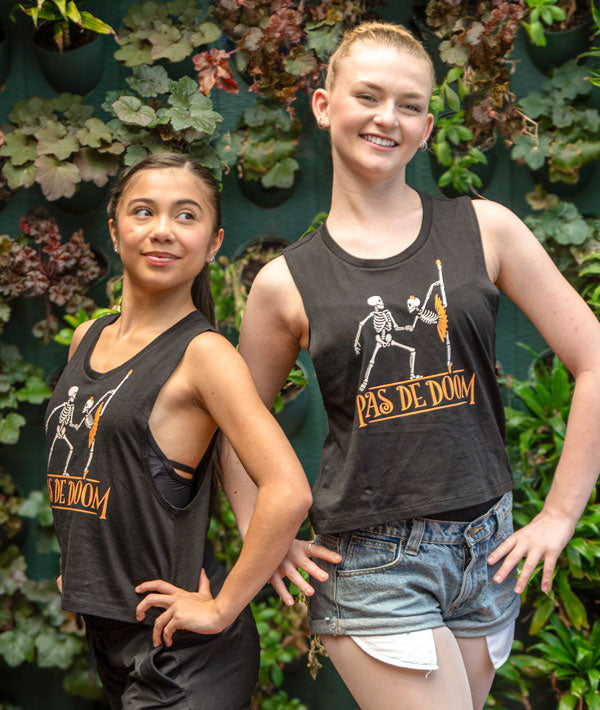 Two dancers wearing black muscle tanks with 'Pas de Doom' design in front of green foliage.