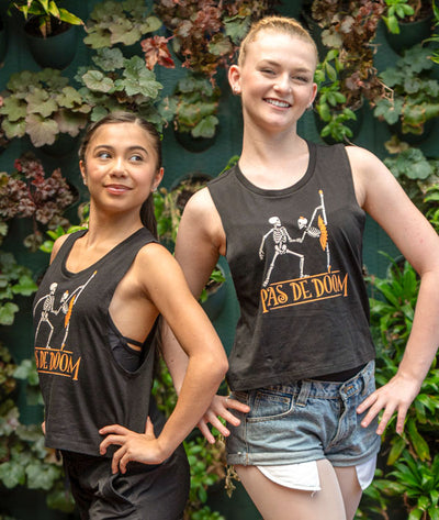 Two dancers wearing black muscle tanks with 'Pas de Doom' design in front of green foliage.