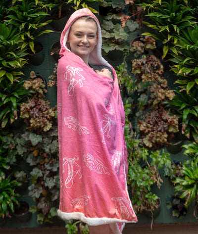 Ballerina wrapped in a pink blanket with white illustrations of pointe shoes and tutus against a green leafy background