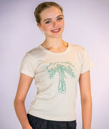 A smiling young ballerina wearing a cream-colored t-shirt with a green graphic of ballet slippers and the cursive text 'pointe your mistletoes'.