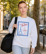 Ballerina walking outside wearing white sweatshirt with French ballet postage stamp graphic carry black dance duffel over her shoulder.