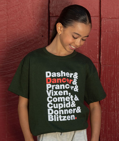 Girl wearing a dark green t-shirt with Christmas-themed reindeer names against a red wood wall.