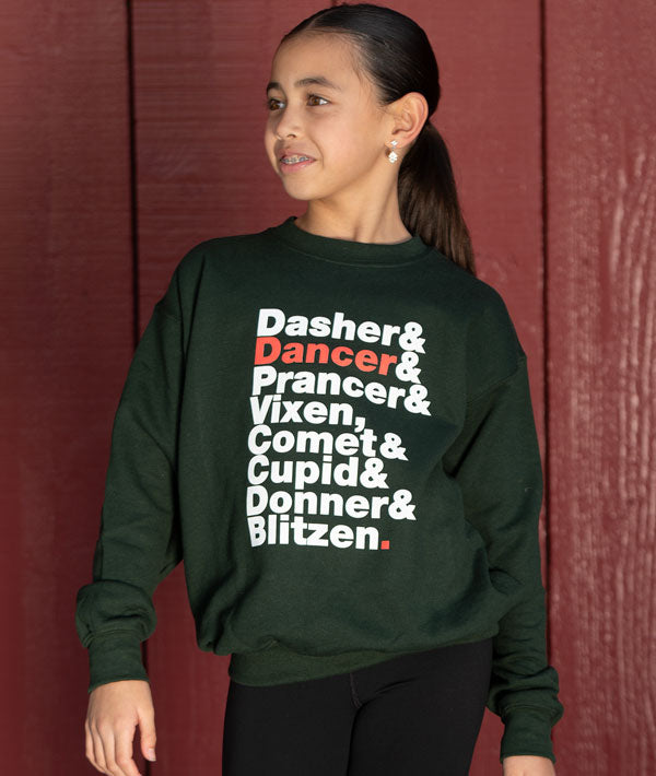 Young dancer wearing a green sweatshirt with Christmas-themed Reindeer names against a red wooden panel background
