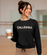 Dancer wearing a black sweatshirt with 'BALLERINA' spelled out as if written with toe tape, holding a coffee cup in a dance studio.