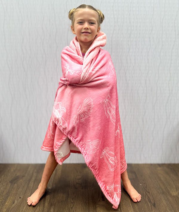 
Young dancer wrapped in a pink blanket decorated with tutus and pointe shoes on a wooden floor and light gray wall background