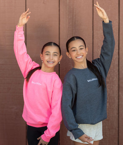Two dancers wearing pink and grey sweatshirts embroidered with 'Ballerina' posing against a brown background.